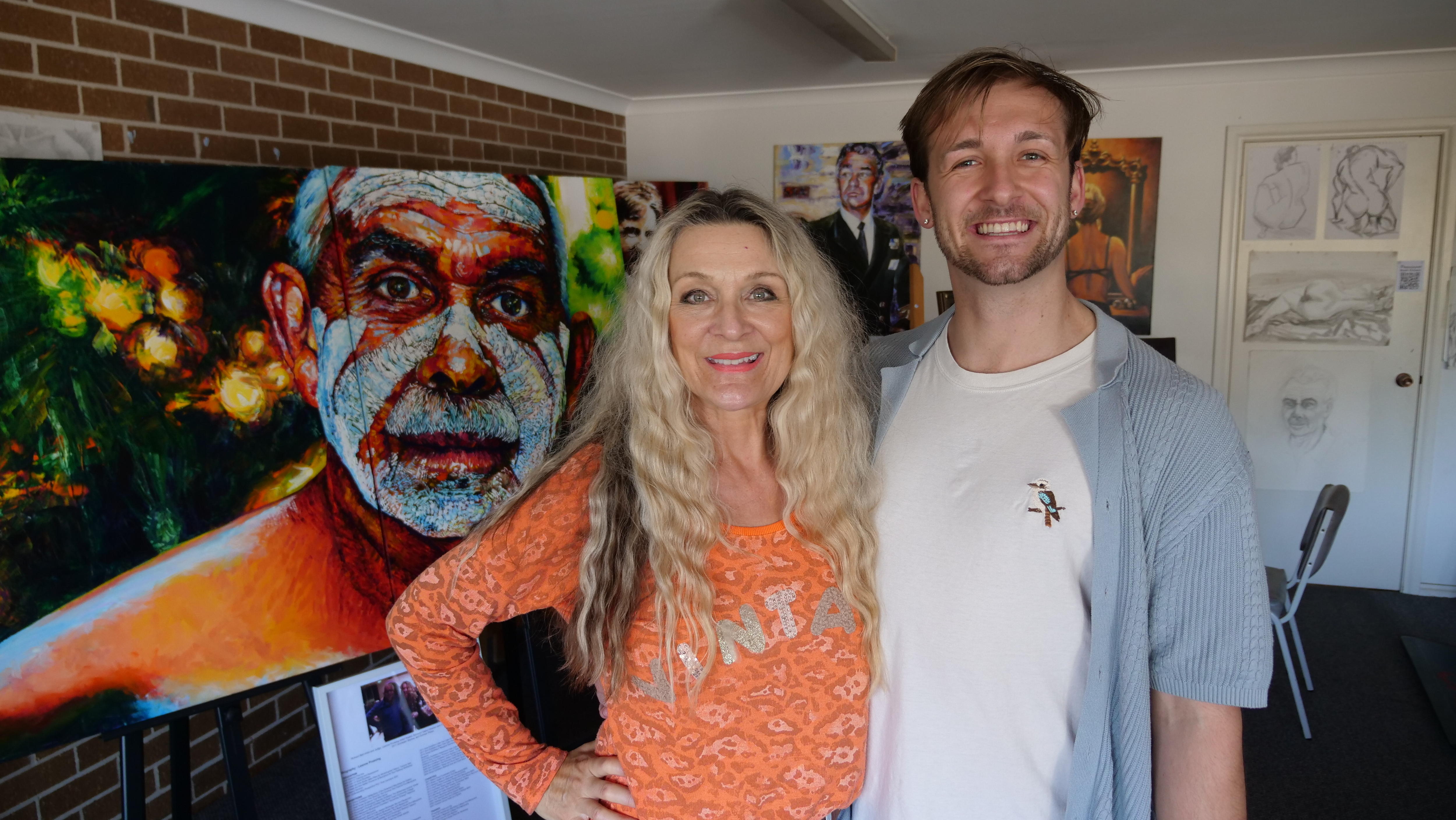 A young man standing with his arm around his mum, who stands in front of her large portrait painting of an Aboriginal man. 
