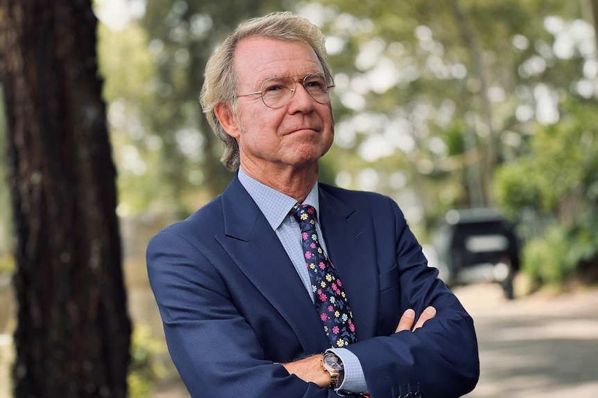 Shane Oliver stands with arms folded, with trees behind him