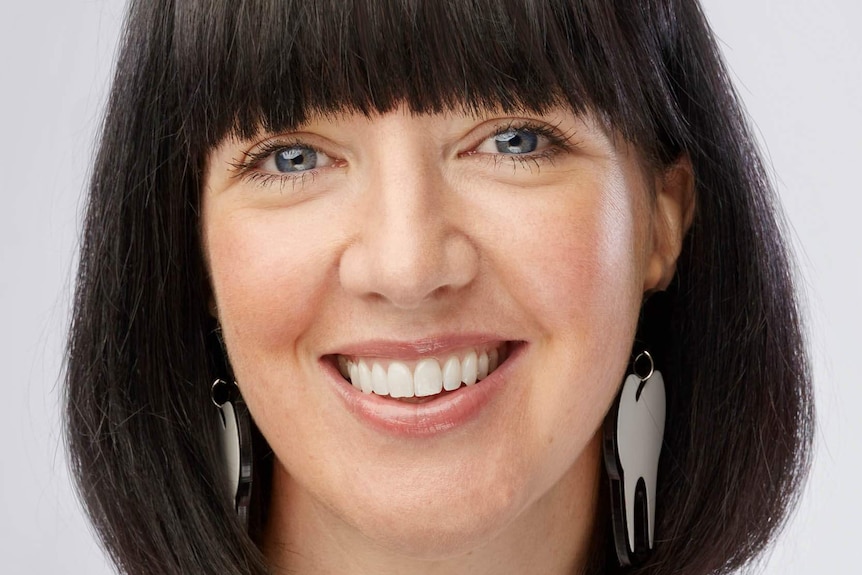 A headshot of a woman smiling, she has a bob of black hair with a thick fringe and wears large earrings shaped as teeth.