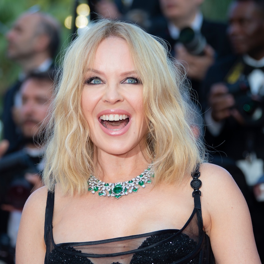Kylie Minogue, wearing a black strappy dress and chunky diamond necklace, smiles on the red carpet