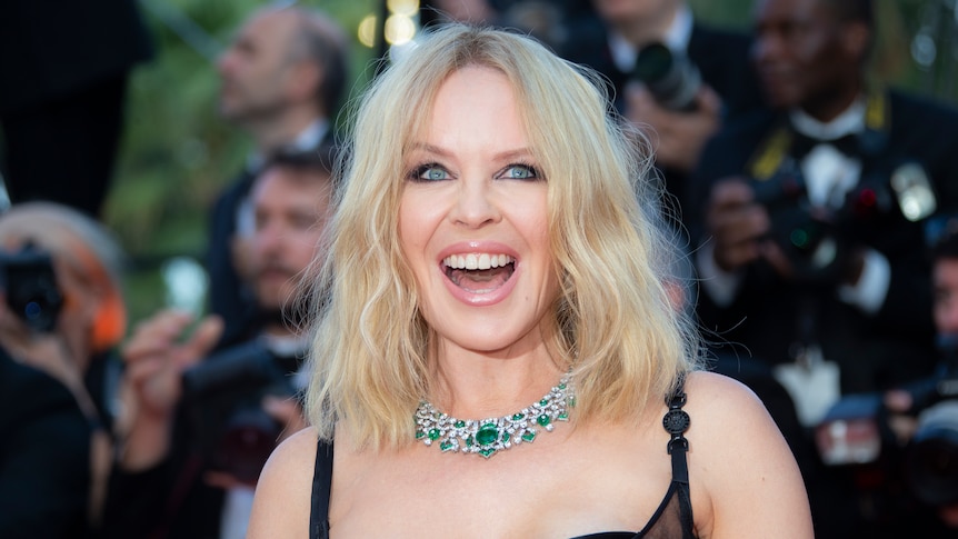 Kylie Minogue, wearing a black strappy dress and chunky diamond necklace, smiles on the red carpet