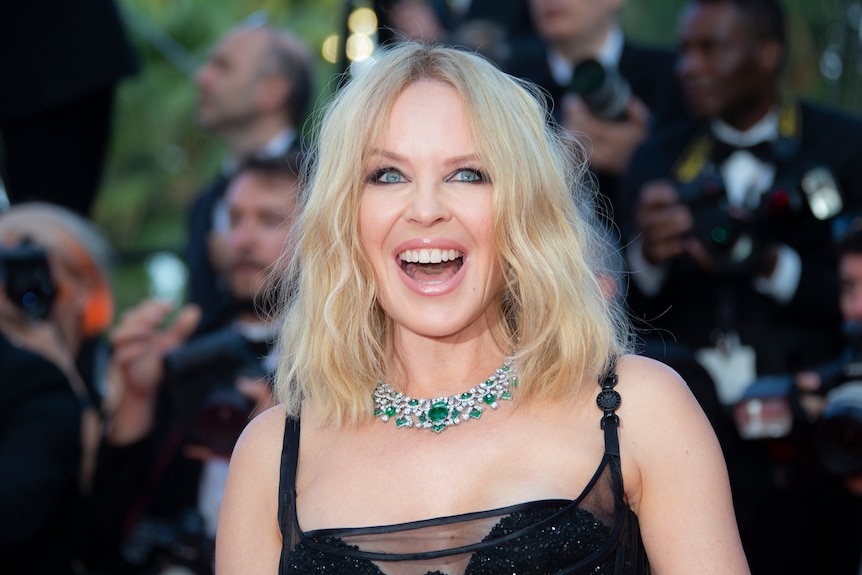 Kylie Minogue, wearing a black strappy dress and chunky diamond necklace, smiles on the red carpet