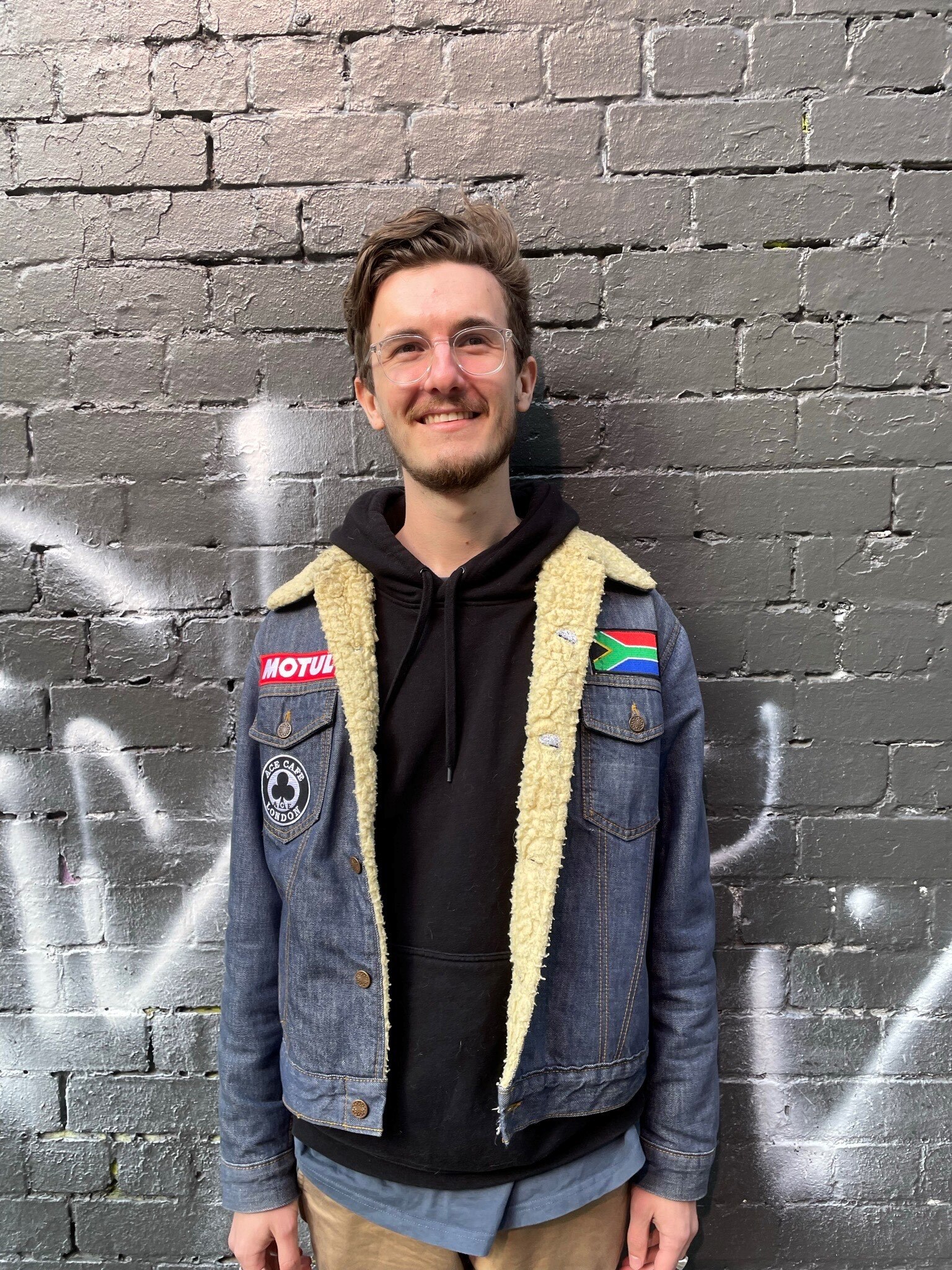 A man smiling while staring at camera, wearing a denim jacket with patches.