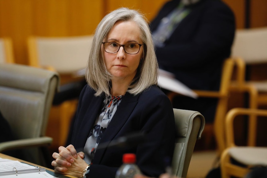 A woman wearing glasses sits with her hands clasped at a bench.