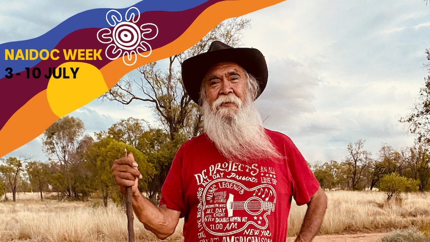 Kamilaroi elder Bob Weatherall standing in the scrub looking at the camera wearing a hat and red t shirt holding a stick