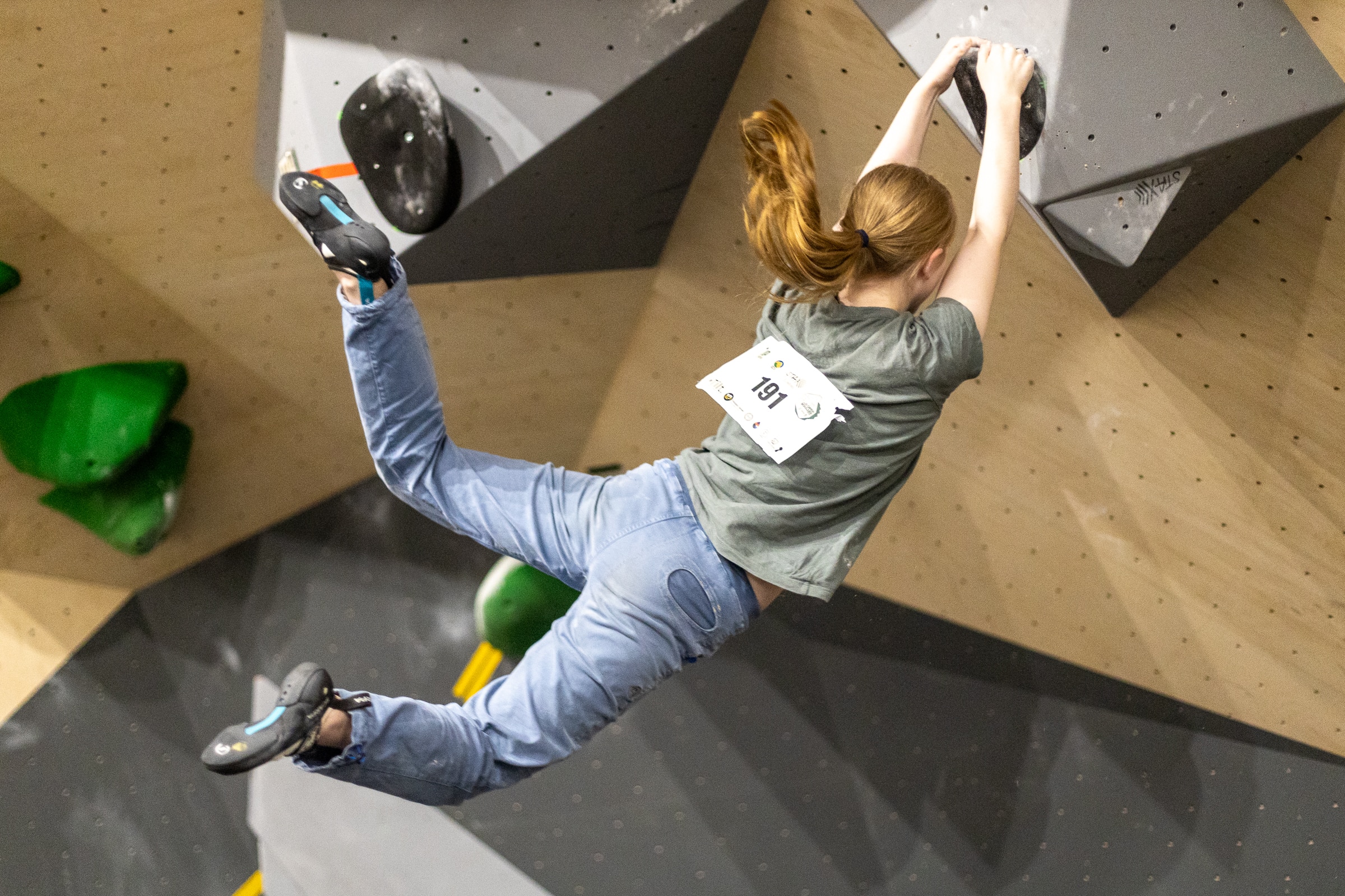 A person with a ponytail is holding onto a rock-climbing wall with legs swinging, suspended.