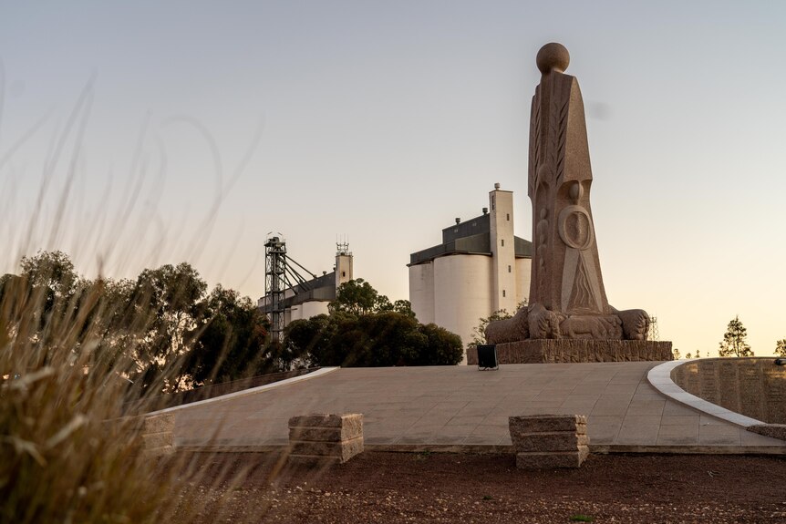 A large stone statue outside with wheat silos in the background at sunset, with long grass in the foreground.