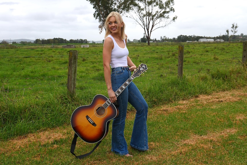 A woman with blonde hair, wearing a white singlet and blue jeans, holding a guitar by her side looking back into a paddock