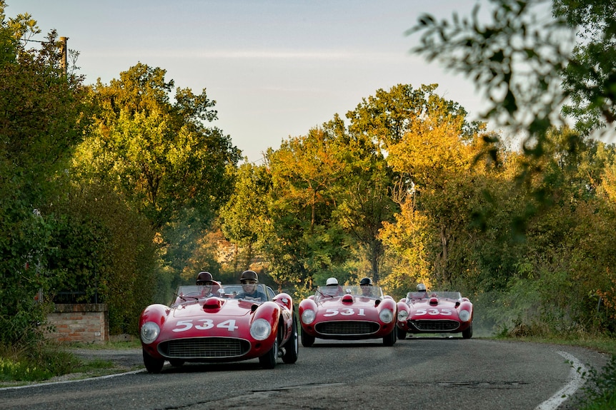 A film still of three red Ferrari cars driving down a road lined by trees