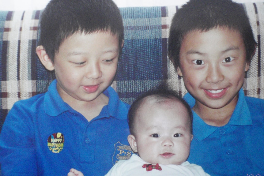 Two young children in school uniforms hold a baby wearing white, who looks to the camera. 