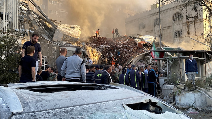 People crowd around a ruined building as smoke rises from it