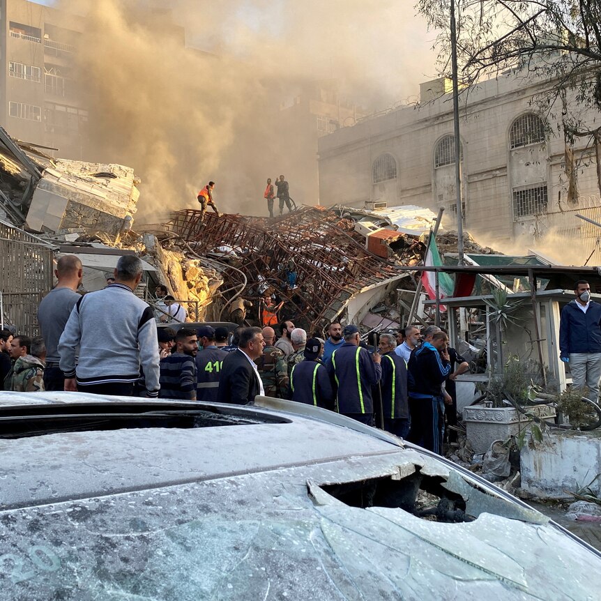 People crowd around a ruined building as smoke rises from it