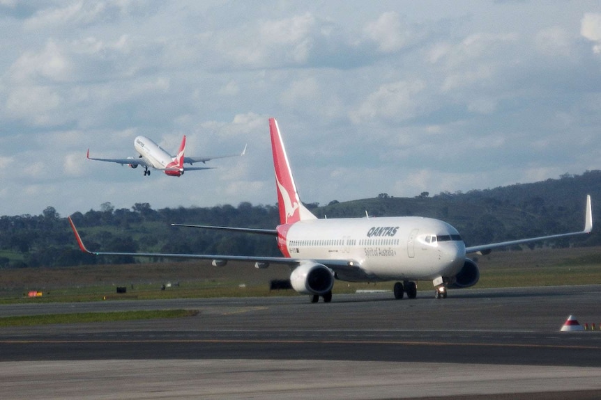 A Qantas plane can be seen taking off as another taxis along the runway.