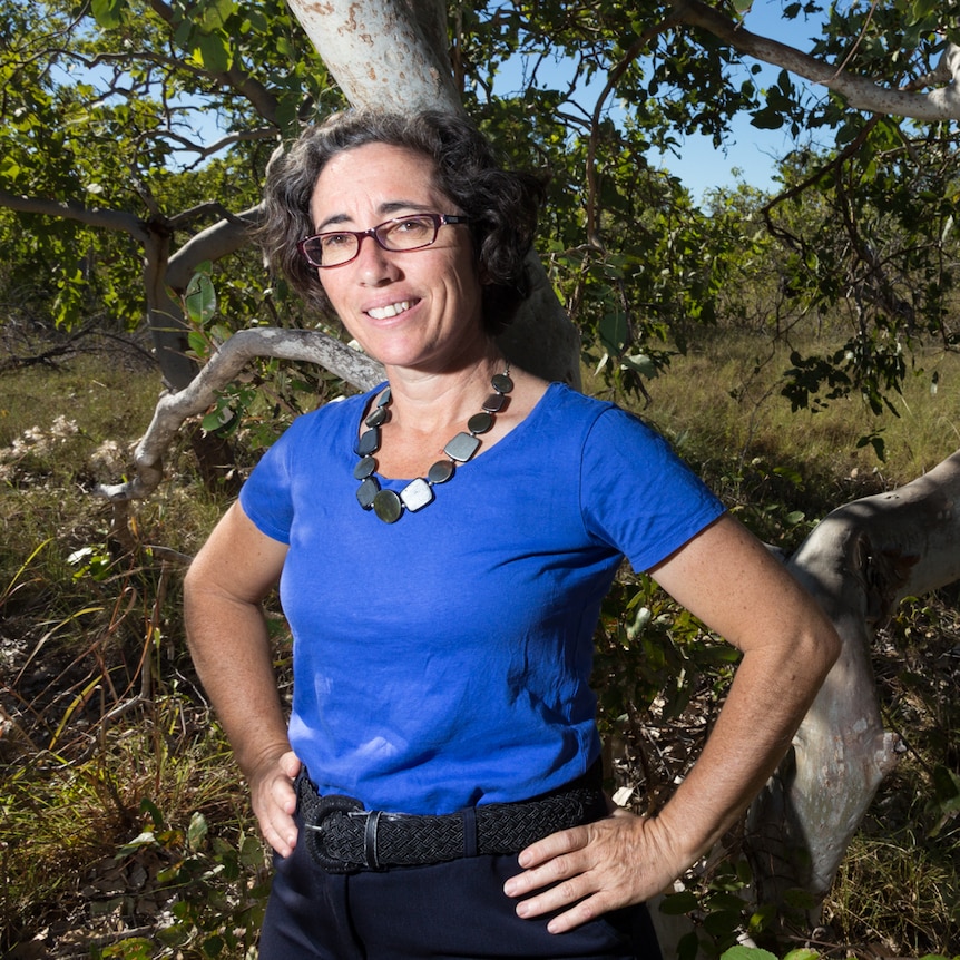 A woman with dark curly hair, wearing glasses and a blue top, standing in front of a gum tree.