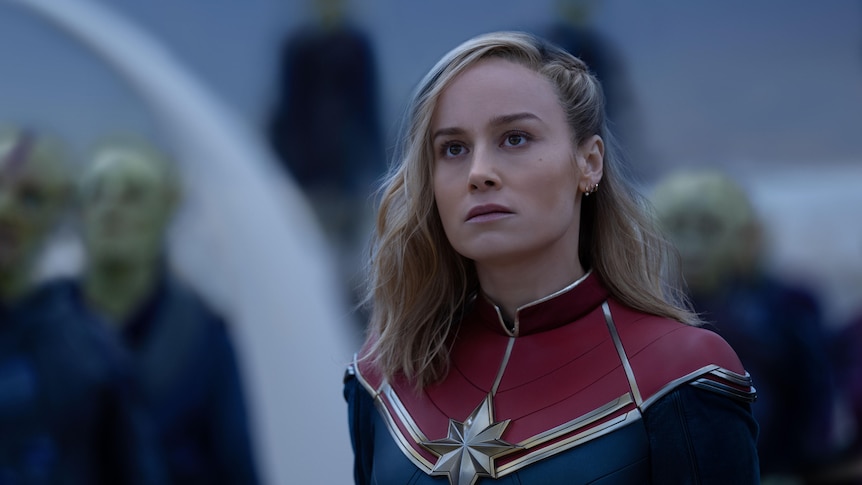 A blonde white woman in her 20s wearing a red and navy superhero costume stares intensely at something off-camera.