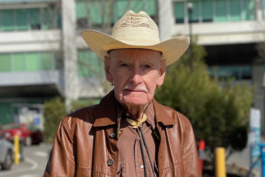 An elderly man wearing a straw cowboy hat and brown leather jacket outside a hospital