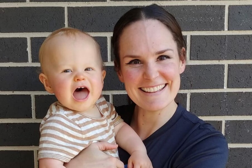 Kelly Wilson in blue t-shirt smiles and holds a blonde baby in cream striped t-shirt, has mouth open. 