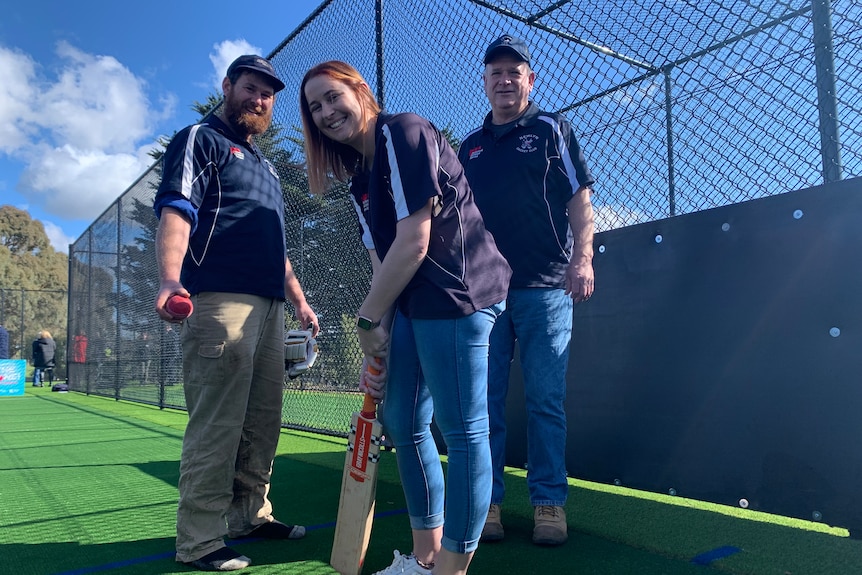 Three people stand in a cricket nets, one holding a cricket bat and one a ball. 