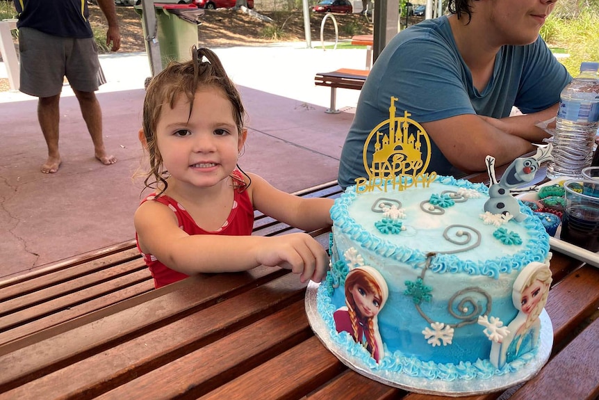 A little girl still wet from swimming sitting down at a picnic table with a blue birthday cake