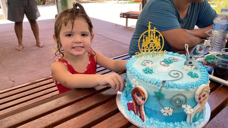 A little girl still wet from swimming sitting down at a picnic table with a blue birthday cake