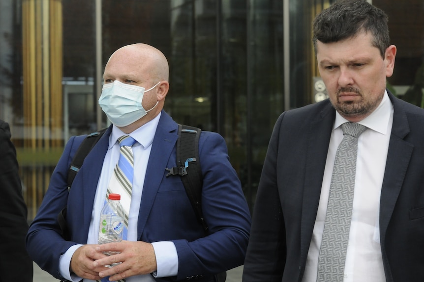 A short, bald man in a white face mask walks beside a man in a black suit.