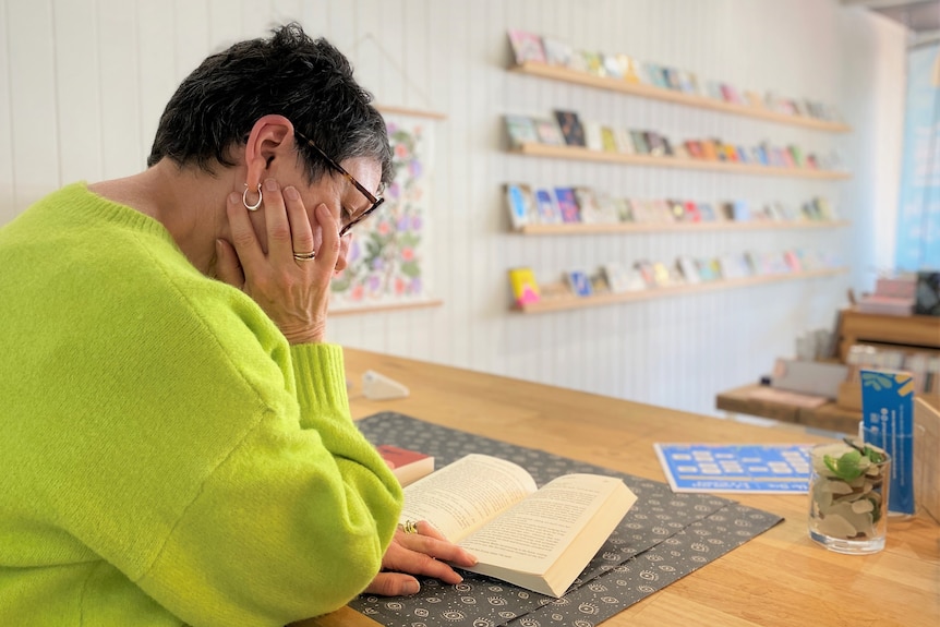 A woman with short dark hair and a bright green jumper stands at a counter with an open book, reading.