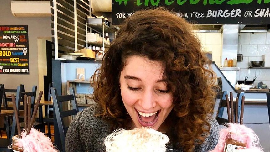 A young woman smiles as she sits in a food shop with three large, colourful milkshakes that are adorned with extra treats.