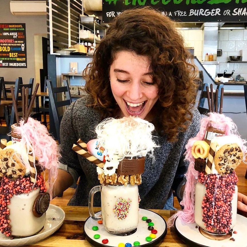 A young woman smiles as she sits in a food shop with three large, colourful milkshakes that are adorned with extra treats.