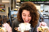 A young woman smiles as she sits in a food shop with three large, colourful milkshakes that are adorned with extra treats.