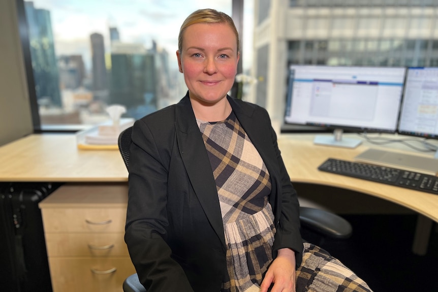 A fair-haired woman in a large-checked dress and dark jacket sits in a CBD office with two computer screens on her desk. 