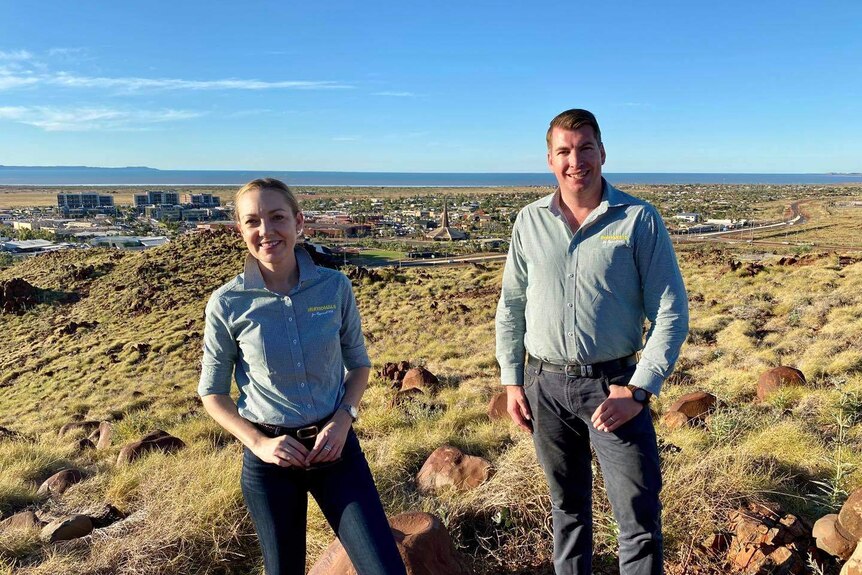 A woman and man in Nationals' shirts, stand smiling on a hill outside the town of Pilbara, with the ocean as backdrop