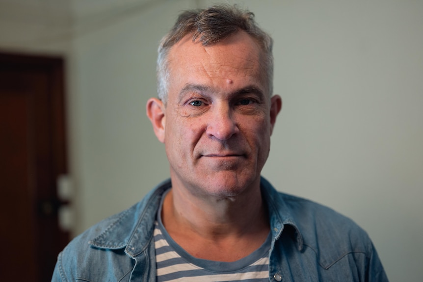 A man looks into a camera, in a close-up portrait. He is standing indoors.