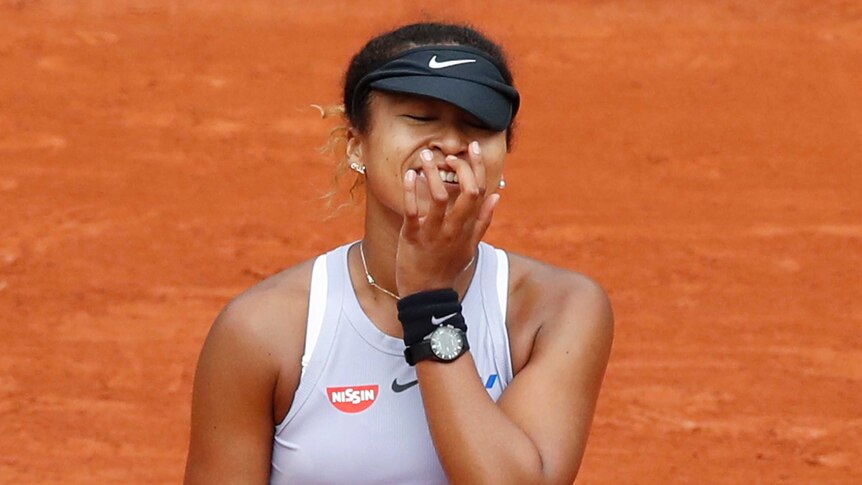 Naomi Osaka holds her hand to her face and leaves her visor cap half pulled down over her forehead.