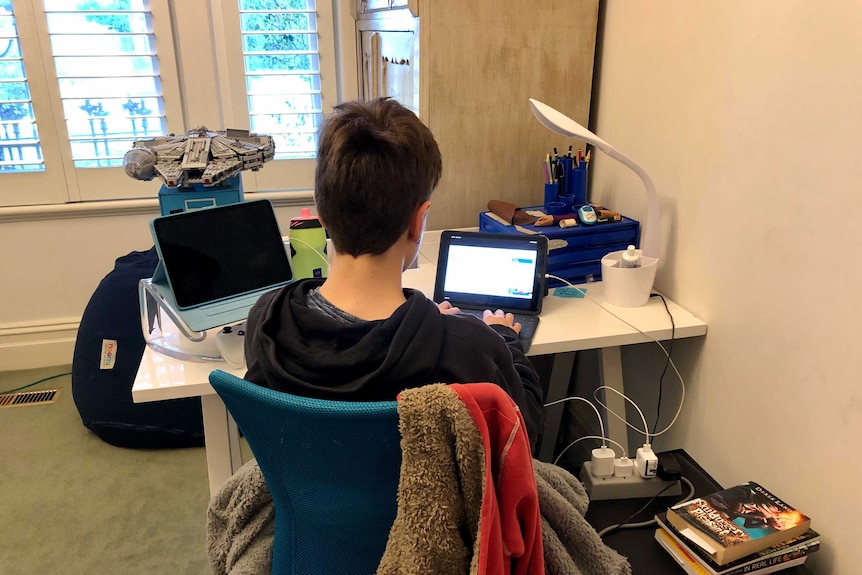 A boy typing at a tablet computer on a desk next to a pile of books.