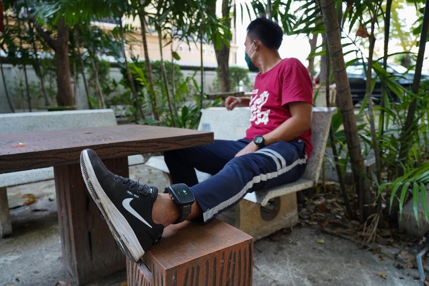 A man faces away from the camera while resting his foot, with a tracking bracelet on his ankle, on a chair