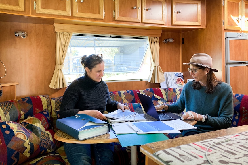 Bec Nicoll and Sarah Thomson sit a table near a window in their caravan, going over documents.