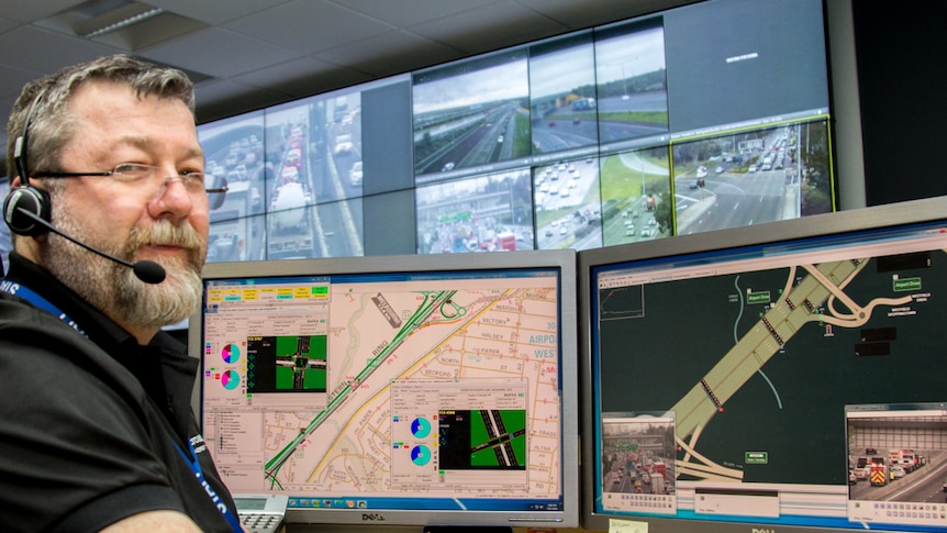 A man sits at a desk with computer screens in front of him, a video wall showing freeways in the background.