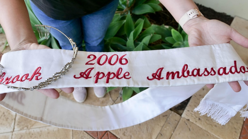 A woman holding a sash that says 2006 Apple Ambassador and a crown. 