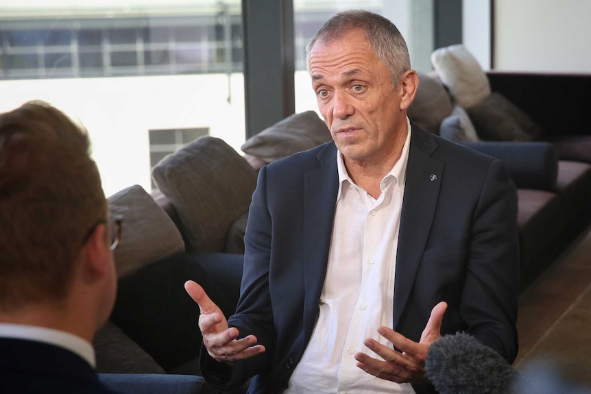 Professor Petit sits on a chair, gesturing with his hands. He wears on open collared shirt.