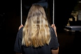 An unidentified teenage girl with blonde hair sits on a swing facing away from the camera.