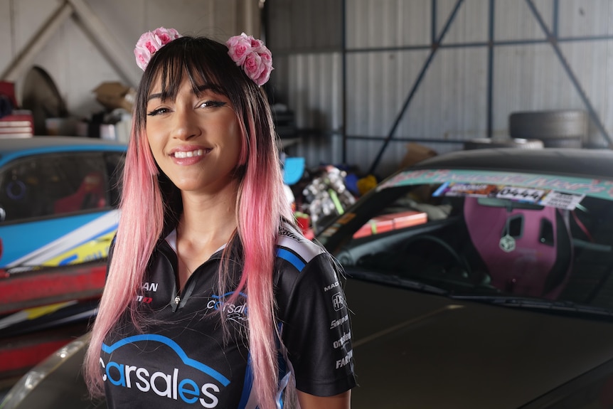 A woman with long pink hair and pink headbands standing in front of a car smiling