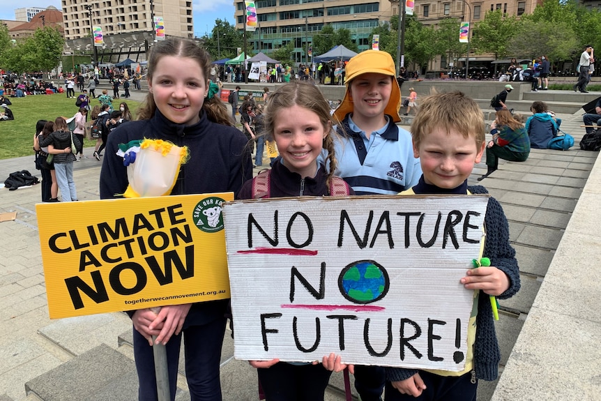 Four children stand together in front of a square. They hold signs that read 'Climate Action Now' and 'No Nature No Future'