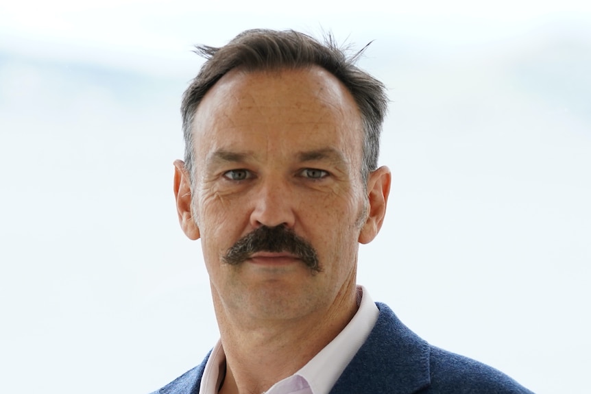 A profile head shot of a white middle-aged male with a short dark hair, greying temples and a brown moustache in a suit.