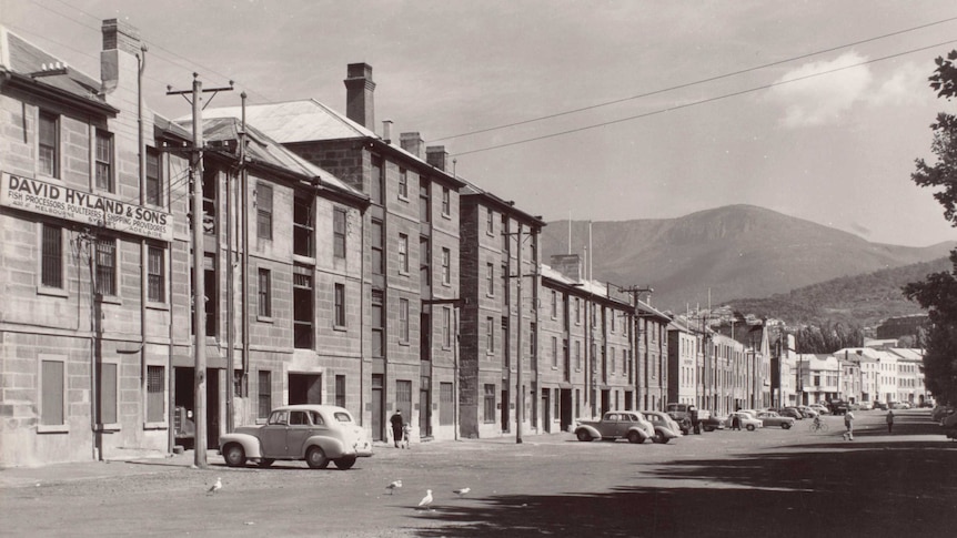 Black and white photograph of Salamanca Place with a dirt carpark and cars parked out the front of the buildings.