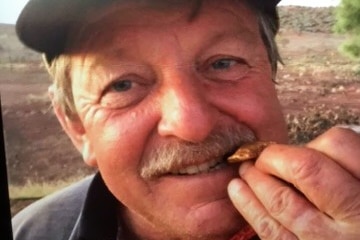 A close-up of a man with a moustache, biting a small gold nugget.