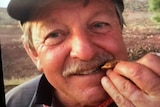 A close-up of a man with a moustache, biting a small gold nugget.
