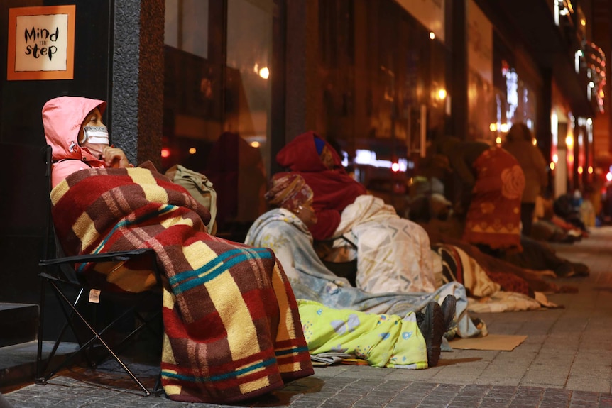Women wearing face masks sit in a queue along an urban road while wrapped up in blankets and coats