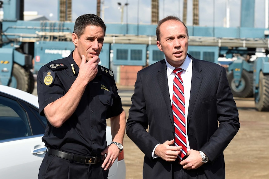 Roman Quaedvlieg stands with one hand on his hip and the other stroking his chin, next to Peter Dutton.