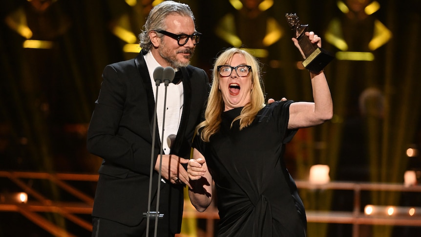A man in his 60s with grey hair and glasses and a woman in her late 50s with blonde hair and glasses hold up an award