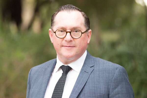 A mid shot of a man wearing a suit and tie and spectacles posing for a photo outdoors.